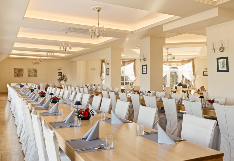 Przestronna sala bankietowa w hotelu z długimi nakrytymi stołami, serwetkami, czerwonymi kwiatami i miękkim oświetleniem.
