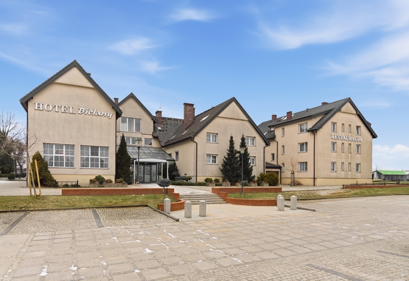 Beżowy hotel z dachami dwuspadowymi, przeszklonym wejściem, zagospodarowanym dziedzińcem i restauracją pod błękitnym niebem.