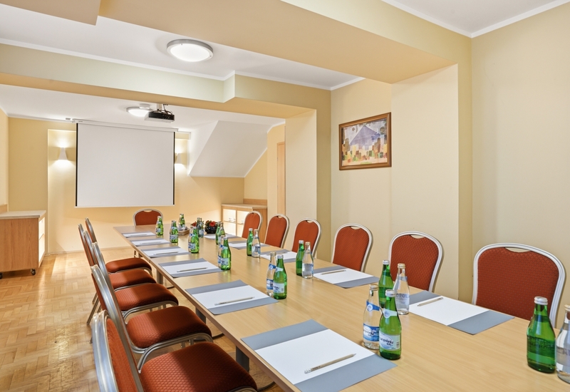 Mała sala konferencyjna z długim stołem, czerwonymi krzesłami, butelkami wody i notatnikami, rzutnik i ekran, ciepłe beżowe ściany