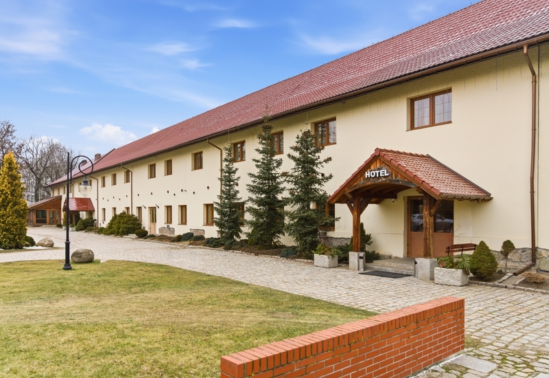 Dwupiętrowy hotel z czerwoną dachówką i drewnianym przedsionkiem z napisem 'HOTEL', brukowany podjazd, iglaste nasadzenia i latarnia