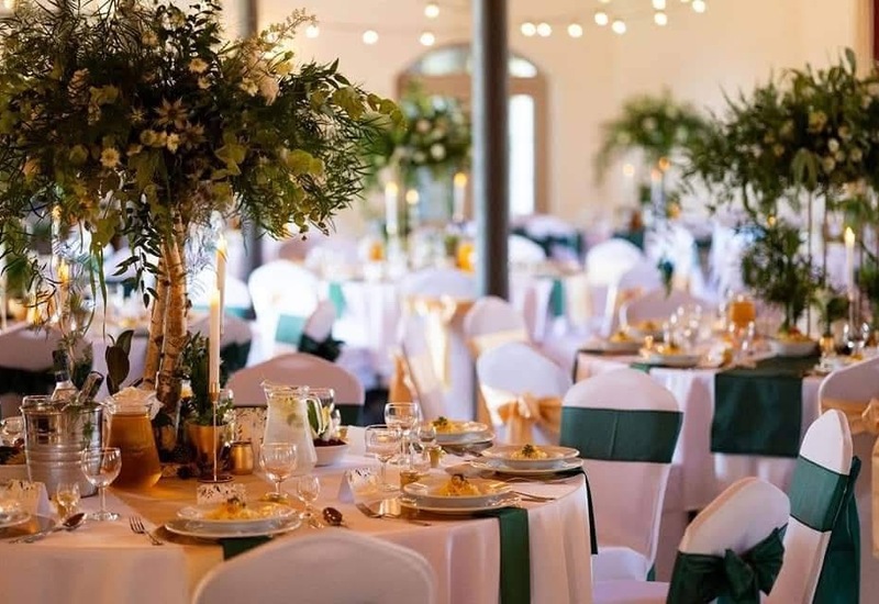 Elegancka sala bankietowa w hotelu przygotowana na wesele: okrągłe stoły w białych obrusach, zielone opaski na krzesłach, wysokie bukiety i świeczki.