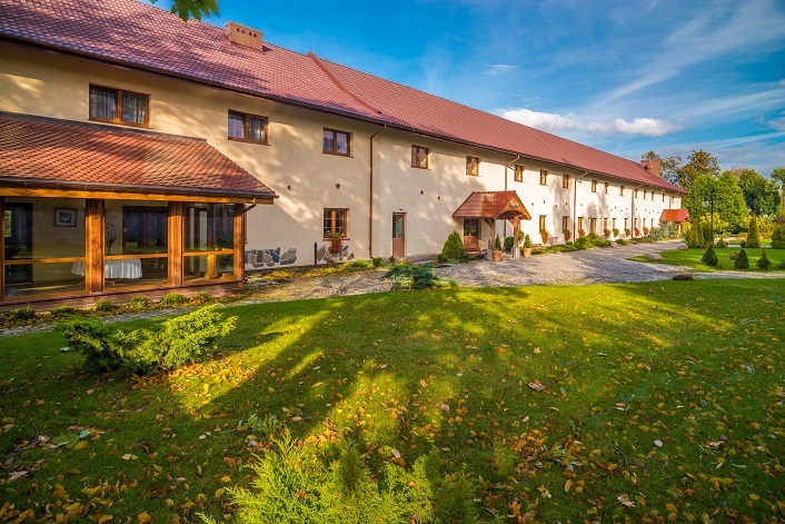 Hotel wiejski z długą kremową elewacją i czerwonym dachem, słoneczny trawnik i krzewy wokół przytulnego wejścia.