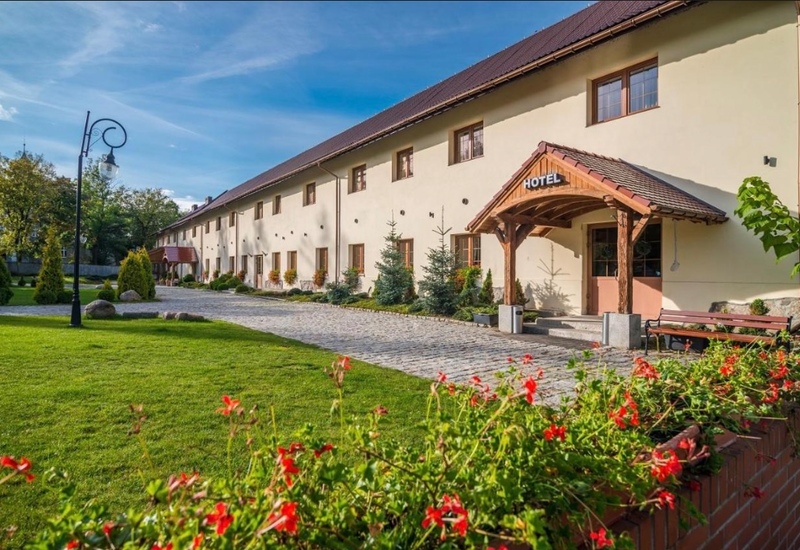 Hotel w stylu wiejskim z drewnianym zadaszonym wejściem, brukowaną ścieżką, zadbaną trawą, czerwonymi kwiatami i ławkami.