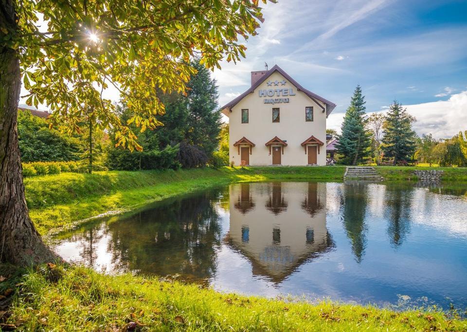 Trzygwiazdkowy hotel wiejski przy spokojnym stawie, z nasłonecznionym trawnikiem i drzewami, idealny na relaks nad wodą.