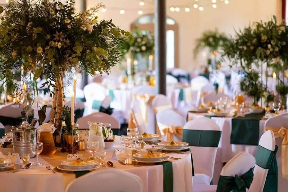 Elegancka sala bankietowa w hotelu przygotowana na wesele: okrągłe stoły w białych obrusach, zielone opaski na krzesłach, wysokie bukiety i świeczki.
