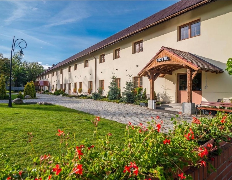 Hotel w stylu wiejskim z drewnianym zadaszonym wejściem, brukowaną ścieżką, zadbaną trawą, czerwonymi kwiatami i ławkami.
