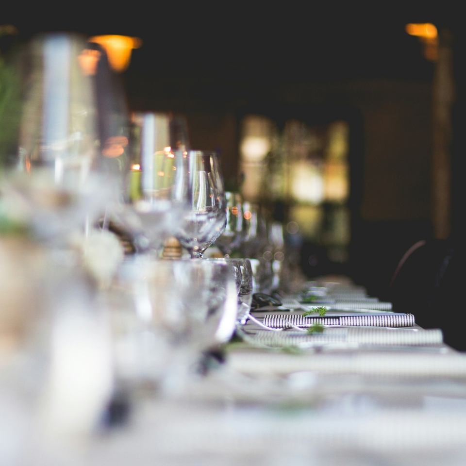 Długi stół w restauracji przygotowany na elegancką kolację, rzędy kieliszków, złożone serwetki i nastrojowe światło świec.