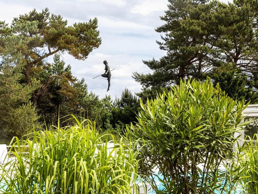 Outdoor pool terrace framed by lush greenery and pines, with a suspended tightrope-walker sculpture above