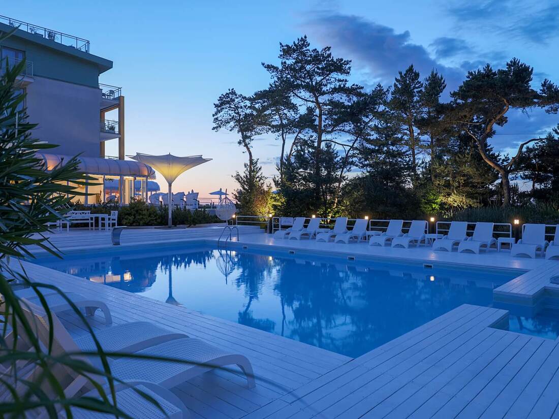 Otwarty basen hotelowy o zmierzchu z białym tarasem i leżakami, podświetlone drzewa i parasole odbijające się w spokojnej wodzie