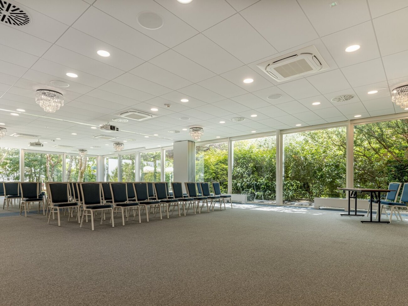 Sala konferencyjna hotelu z rzędami krzeseł, przeszklonymi ścianami z widokiem na ogród, żyrandolami i projektorami.