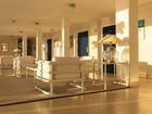 Sunlit hotel lobby corridor with modern white lounge chairs on rugs, glass table lamps, blue guestroom doors and tiled floor