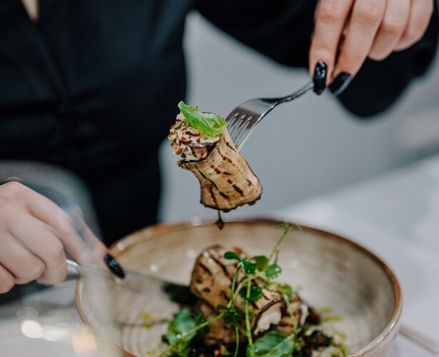 Osoba w eleganckim stroju delektująca się rolowanym daniem z grillowanym bakłażanem i świeżą zieleniną w restauracji hotelowej.