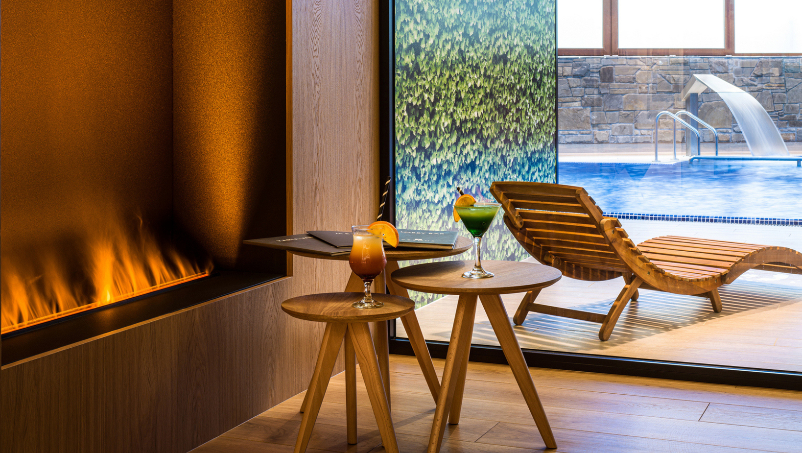 صالة دافئة بجانب موقد خطي، كوكتيلات على طاولات خشبية وكراسي للتشمس بجانب مسبح مع شلال ماء.