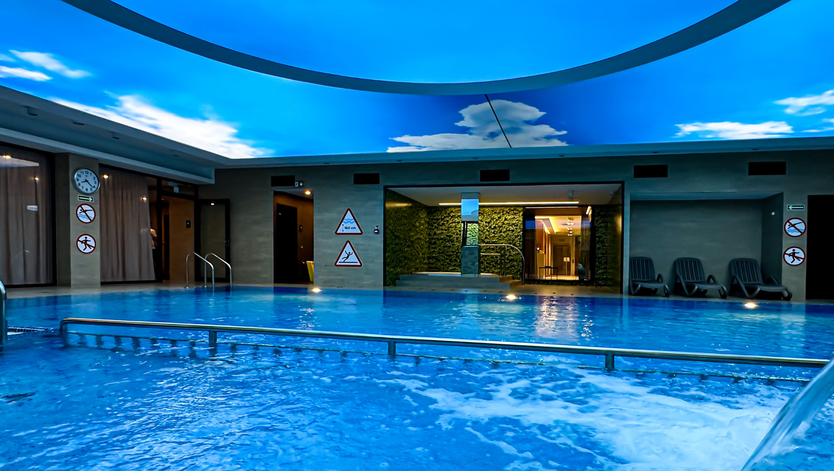 Krytý hotelový bazén pod nebeskou kopulí, bublající trysky, madla, lehátka a vstup do spa se zelenou živou stěnou