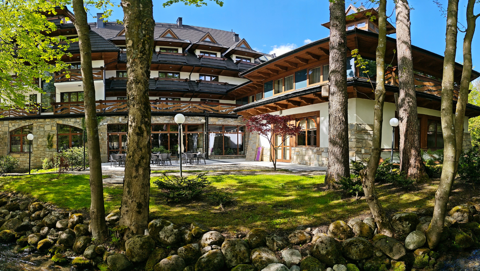 فندق جبلي على طراز فيلا مع شرفات خشبية وواجهة حجرية، تراس مظلل بمقاعد بجانب جدول ماء.