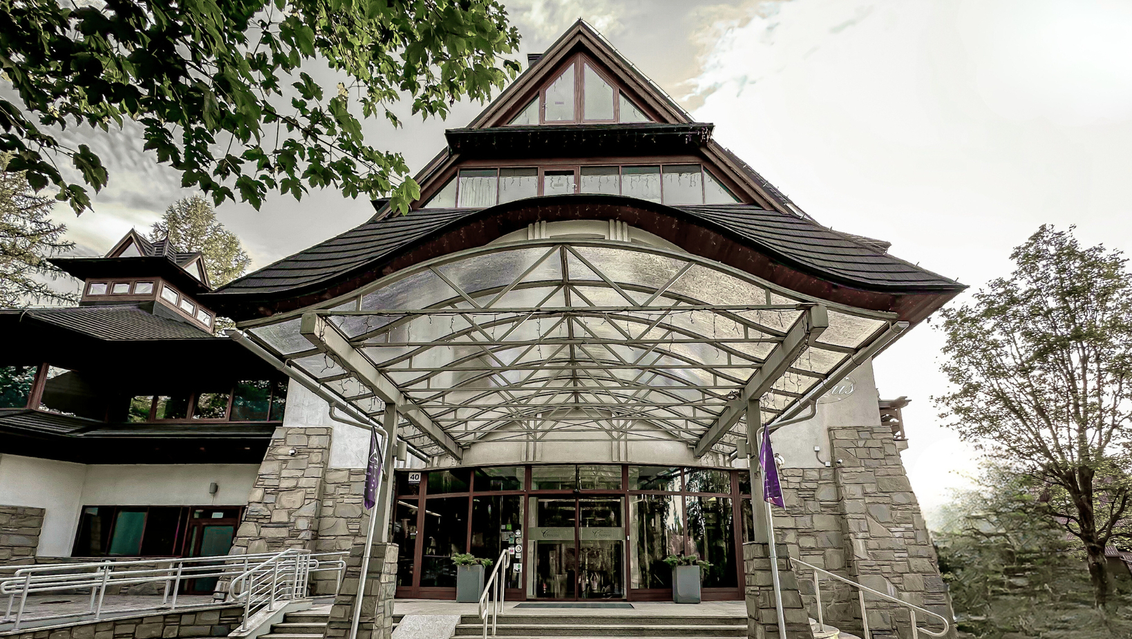 مدخل فندق بطراز جبلي مع مظلة زجاجية وأعمدة حجرية وأبواب زجاجية وممر لذوي الاحتياجات ونباتات مزروعة.