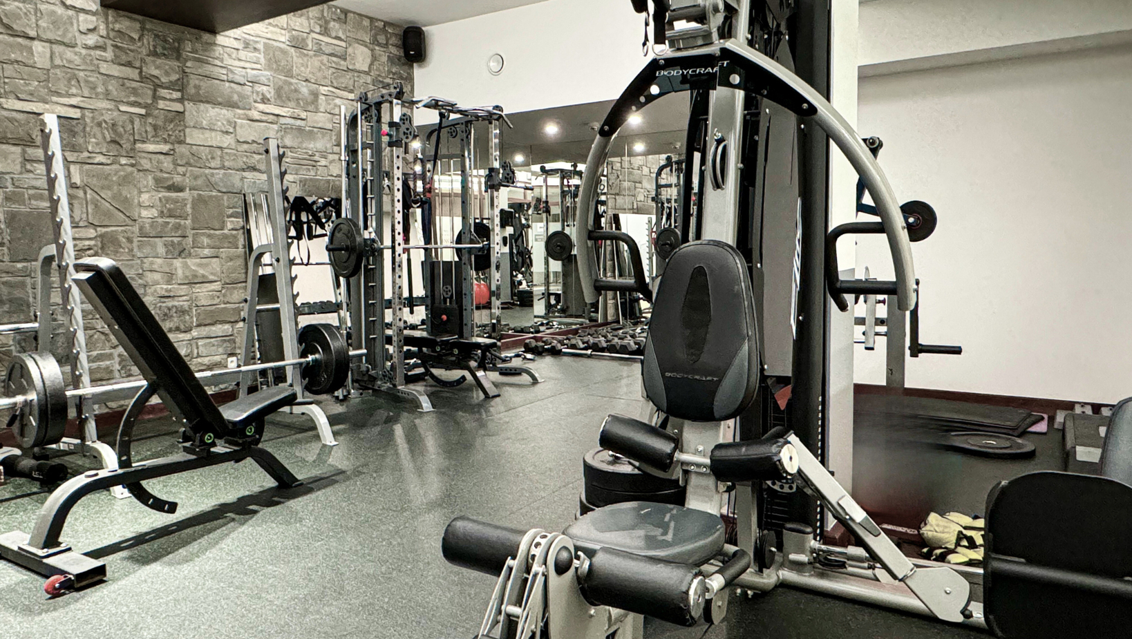Hotelové fitness s viacstanicovým strojom, lavičkou na bench press, voľnými činkami a zrkadlovou kamennou stenou