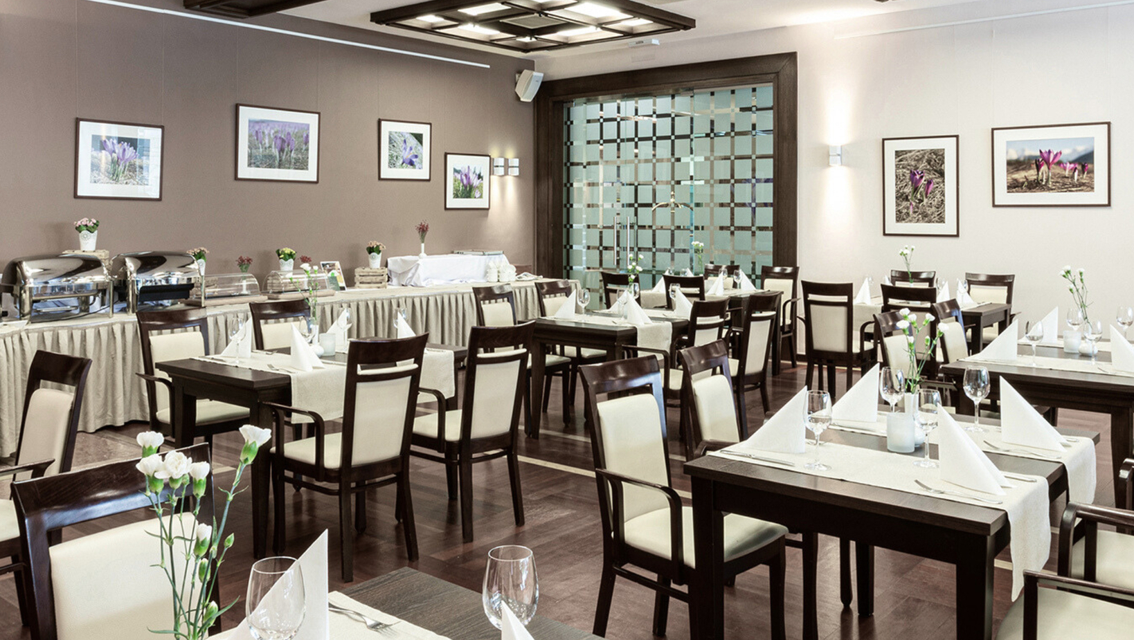 قاعة طعام فندقية راقية بأثاث خشبي داكن وكراسي كريمية، طاولات مُعَدَّة بمناشف مطويّة وكؤوس، ومزهريات زهور وبوفيه بسخانات.