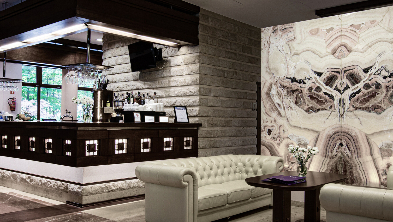 Hotelový salónik s krémovou prešívanou koženou pohovkou, tmavým barom s pohármi a alkoholom, podsvietený mramorový panel a čerstvé kvety