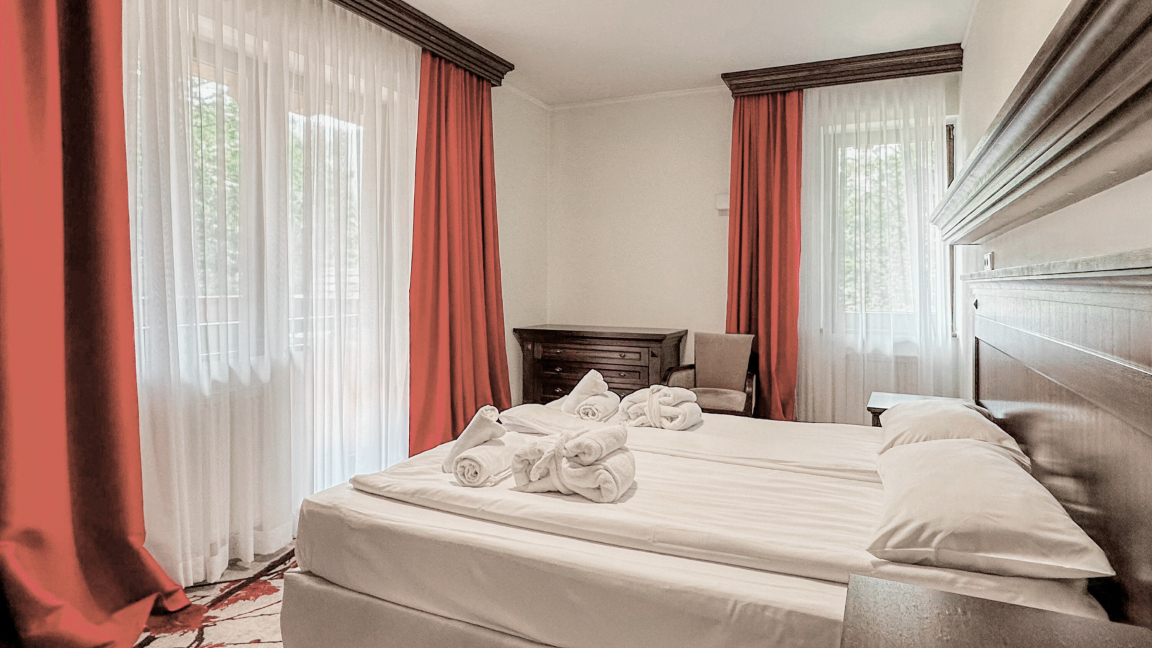 Svetlá klasická hotelová izba s dvoma posteľami spojenými dokopy, bielym posteľným prádlom, navinutými županmi na posteli, dreveným nábytkom a červenými závesmi