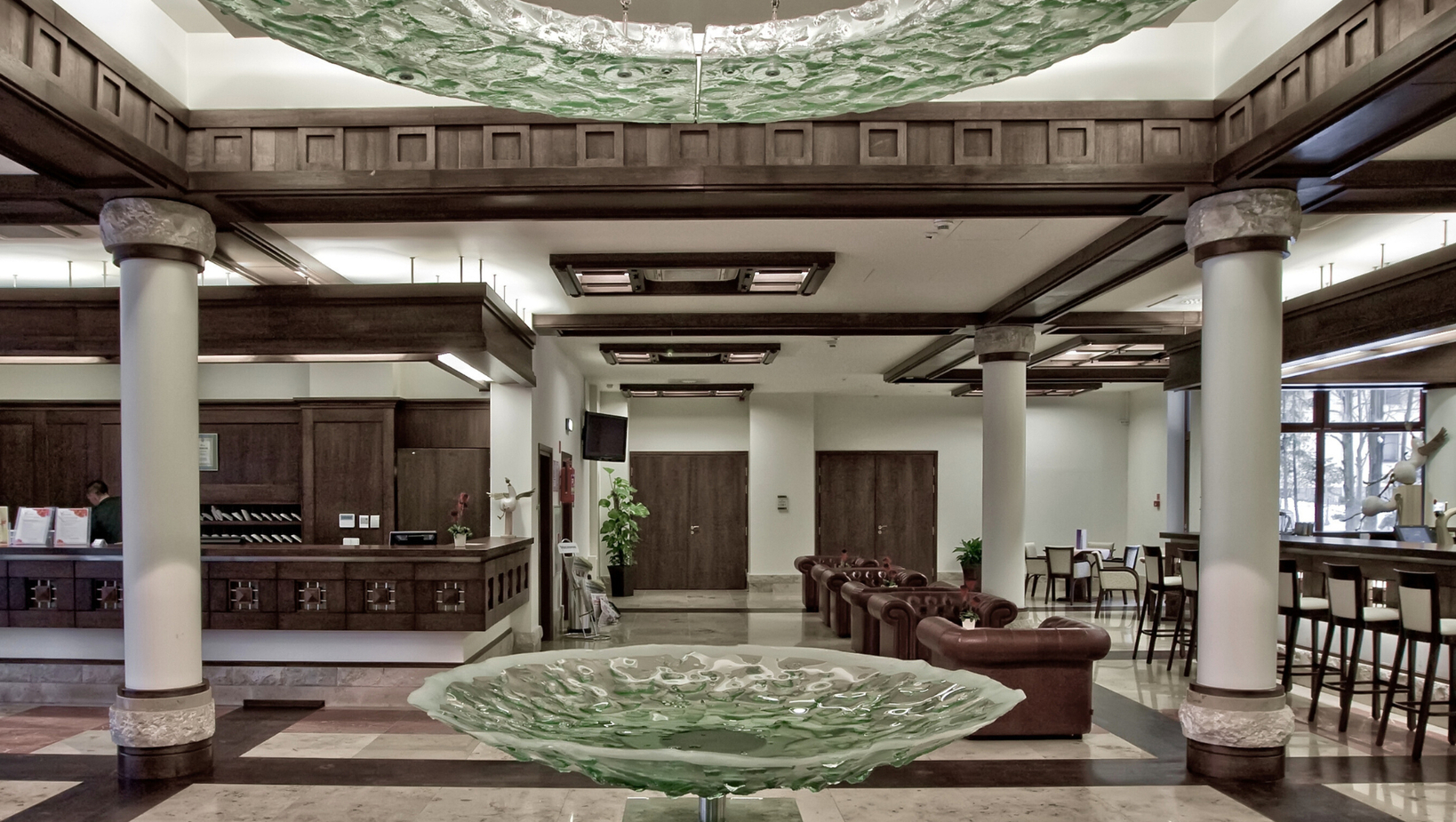 Priestranné hotelové lobby s drevenými trámy, mramorovou podlahou, zelenou sklenenou plastikou, koženými sedačkami, recepciou a barom.