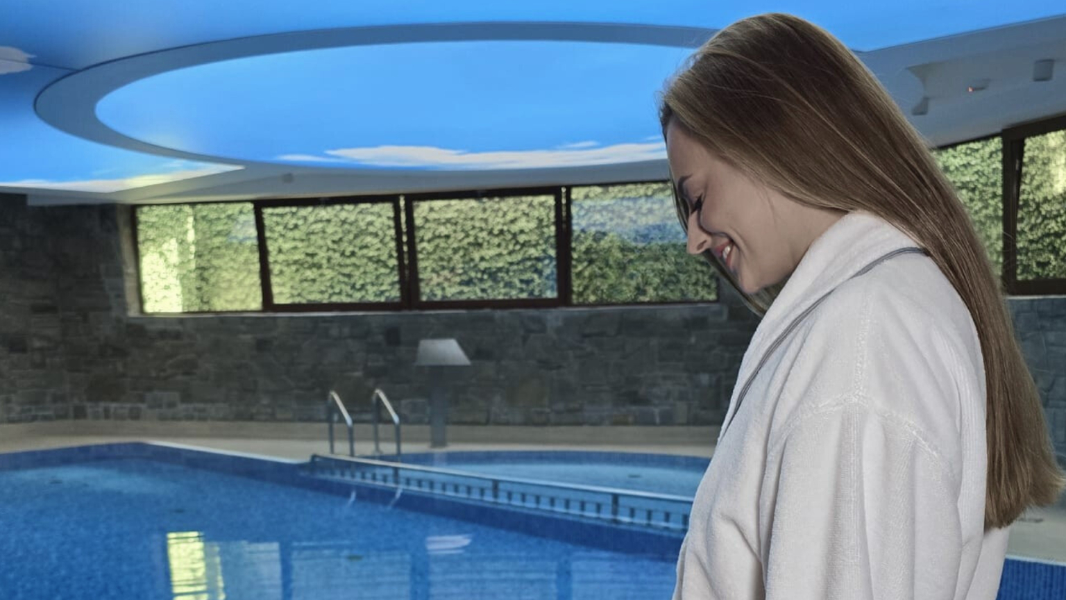 Usmívající se žena v bílém županu u klidného krytého hotelového bazénu s žebříkem, kamennými zdmi a stropem s motivem oblohy.