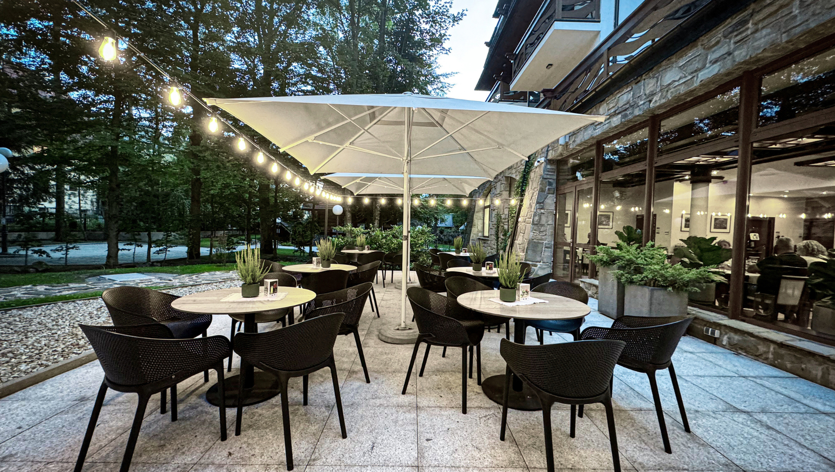 Vonkajšia hotelová terasa s okrúhlymi stolmi, prútenými stoličkami a veľkým bielym slnečníkom, reťazovým osvetlením, kamennou fasádou a stromami pri súmraku