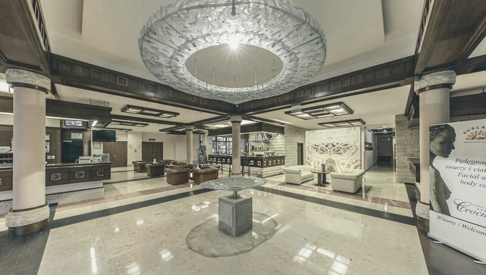 ردهة فندق واسعة بأرضية رخامية، طاولة زخرفية في الوسط، ثريا سقفية دائرية، أرائك جلدية، مكتب استقبال وبار.