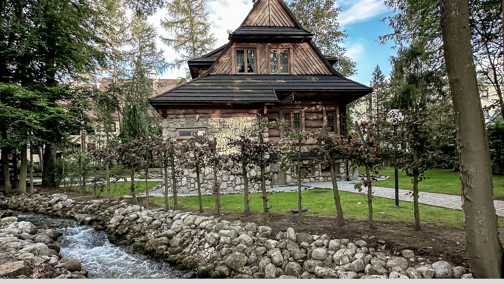 Przytulna górska chata z kamiennym parterem i drewnianym piętrem, ogród nad strumieniem z kamiennym brzegiem i zacienioną ścieżką.