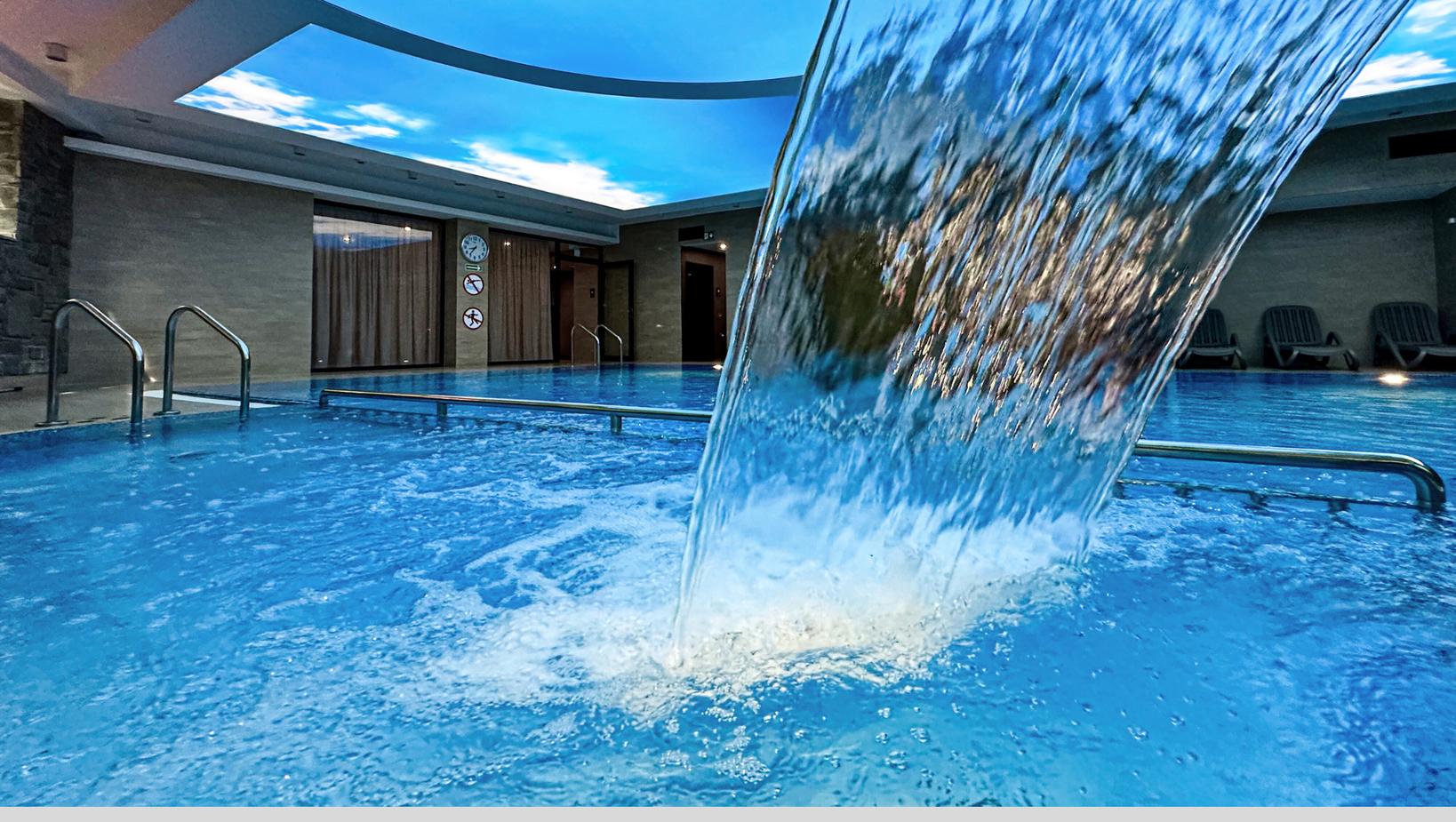 Vnitřní hotelový lázeňský bazén s vodní kaskádou, světlíkem s modrou oblohou, nerezovými madly a lehátky.