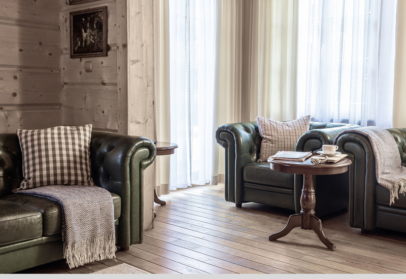 Útulný horský salónik so zelenými koženými pohovkami Chesterfield, kockovanými vankúšmi, drevenou podlahou a stolíkom s šálkou a časopisom.