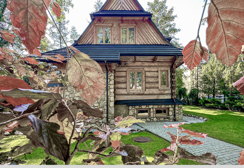 Malebná drevená chata na kamennom podstavci so strmou strechou, dláždenou cestou a upraveným trávnikom, rámovaná červenými listami.