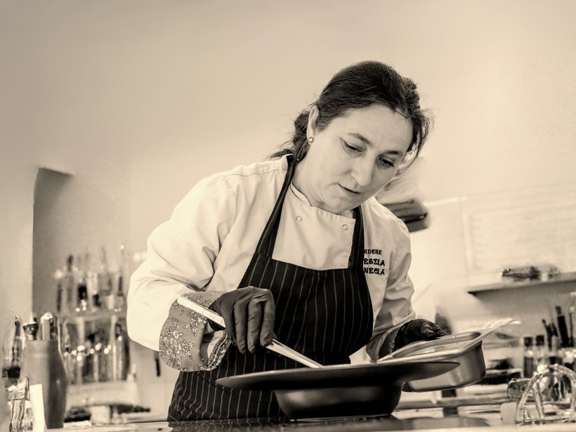 Kuchárka v zástere a rukaviciach upravuje jedlo v profesionálnej kuchyni, sústredená na precízne servírovanie.