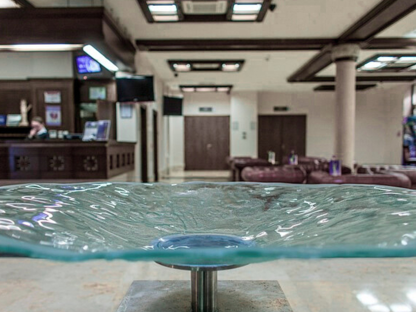 Sklenená miska-fontána na mramorovom podstavci v hotelovej hale; recepcia a kožené sedenie s teplými drevenými akcentmi.