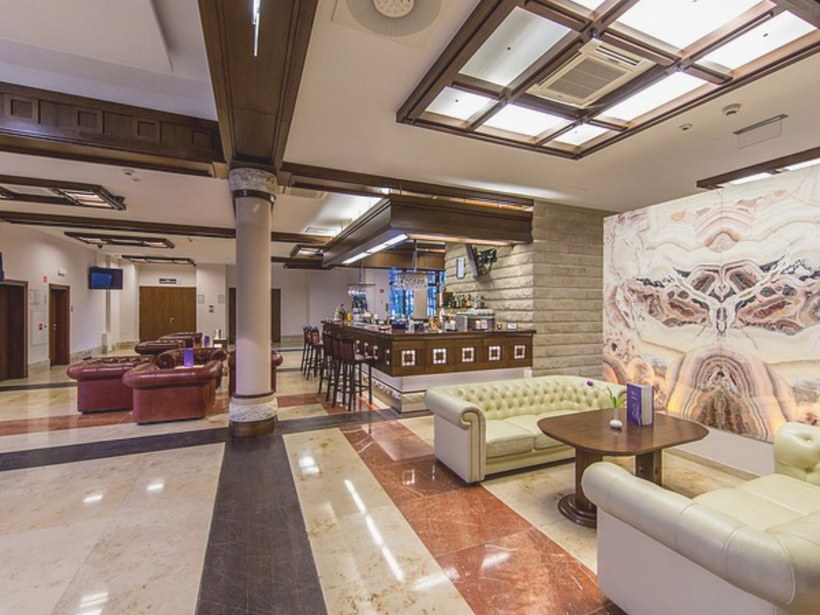 Priestranné hotelové lobby s mramorovou podlahou, prešívanými krémovými sedačkami, drevenými trámami, barom a podsvietenou kamennou stenou.