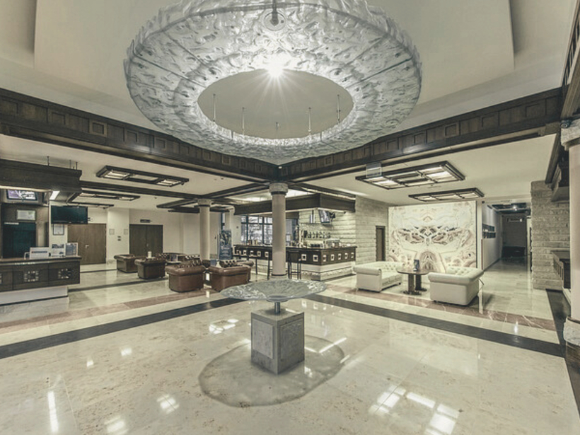 Priestranné hotelové lobby s mramorovou podlahou, centrálnym dekoratívnym stolom, zdobeným kruhovým stropným svetlom, koženými sedačkami, recepciou a barom.