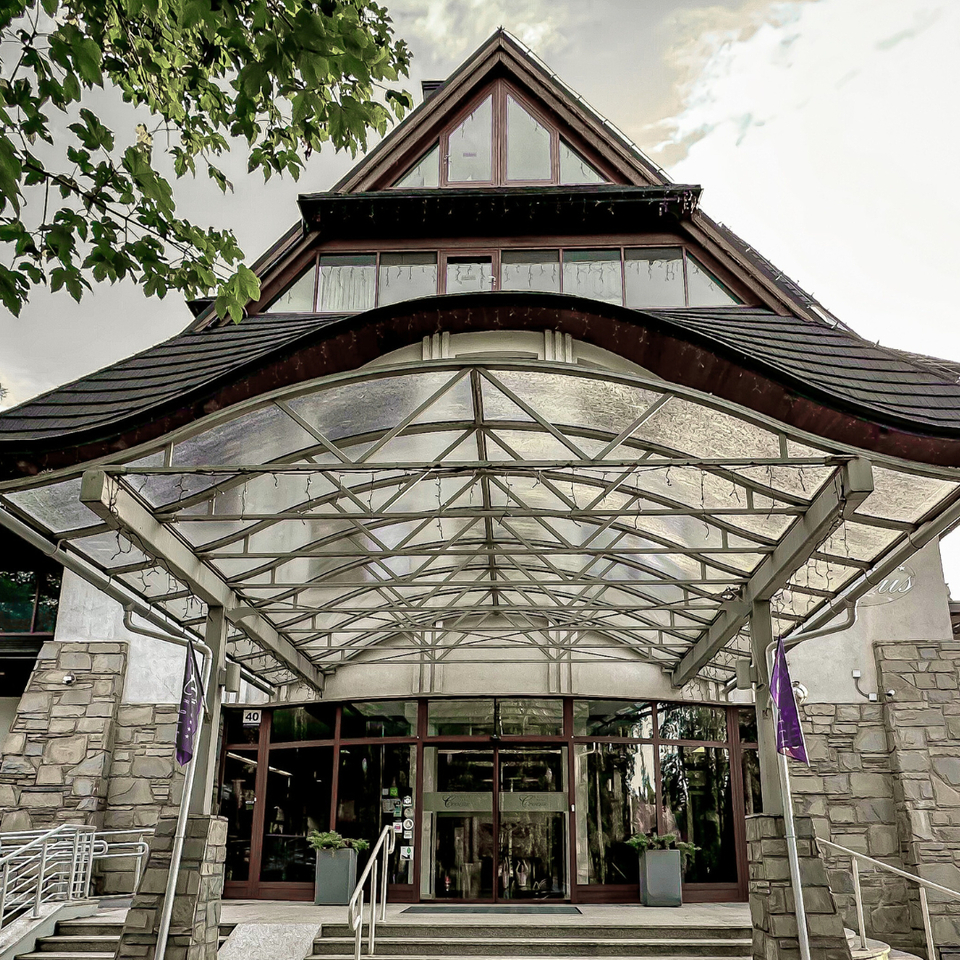 Vstup do hotela v alpskom štýle s preskleným prístreškom, kamennými piliermi, sklenenými dverami, bezbariérovým nájazdom a kvetináčmi.