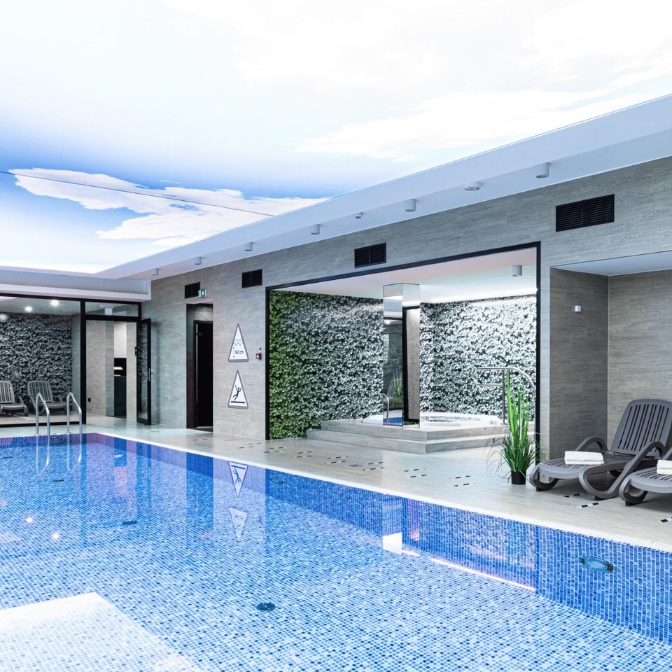 Vnútorný hotelový bazén s mozaikovou dlažbou, stropom s motívom oblohy, ležadlami s uterákmi a vírivkou pri zelenej stene.