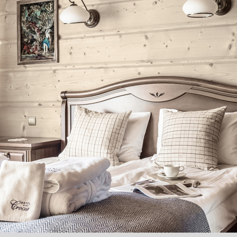 Przytulny pokój z drewnianymi ścianami, łóżkiem dwuosobowym, kratkowymi poduszkami, lampami, złożonymi ręcznikami 'Hotel Crocus', filiżanką i magazynem