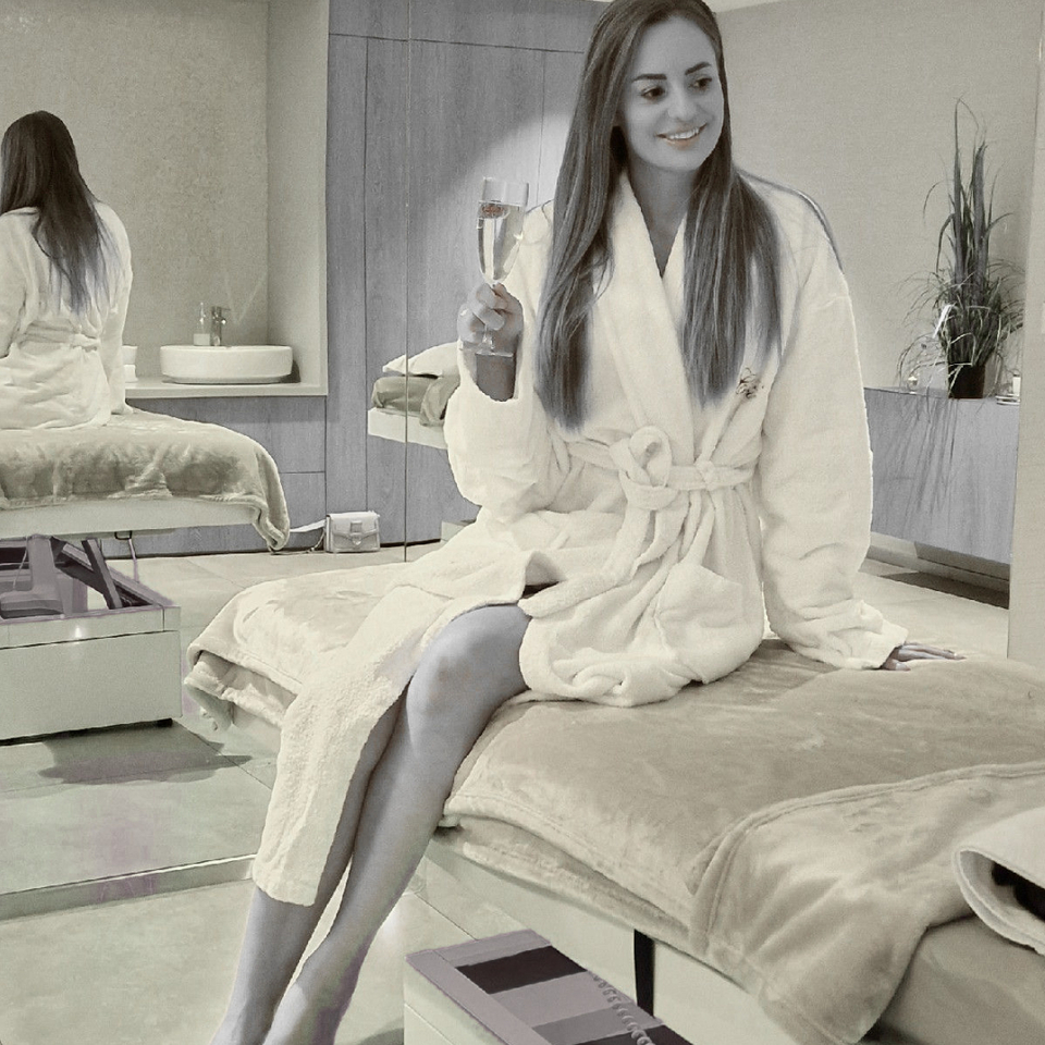 امرأة برداء فخم تحمل كأس شمبانيا جالسة على سرير علاج في غرفة سبا هادئة مضاءة بالشموع