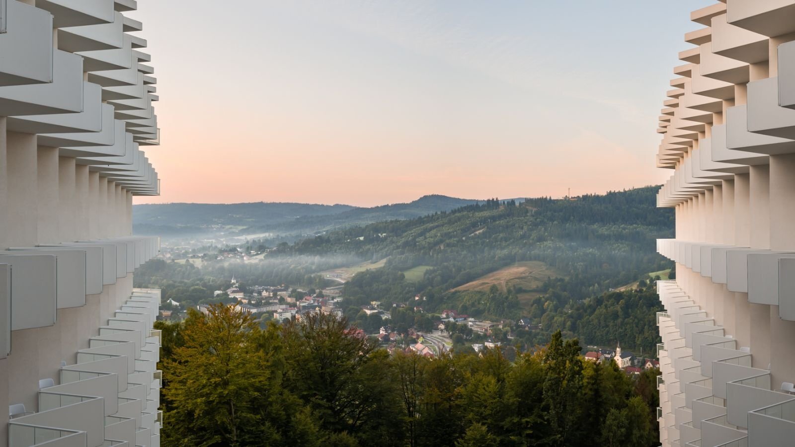 Gestaffelte weiße Hotelbalkone rahmen ein nebliges Tal mit Ortschaft und bewaldeten Hügeln unter zartem Pastellhimmel.