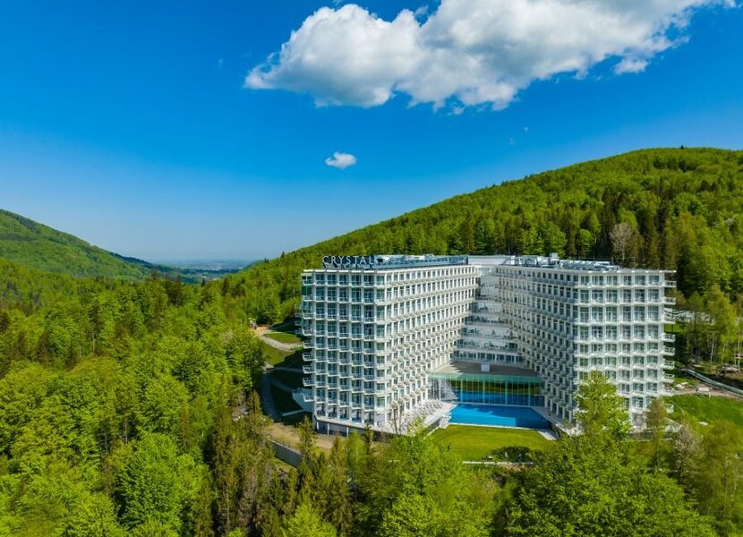 Nowoczesny wielopiętrowy hotel z balkonami i odkrytym basenem, położony w zielonych, zalesionych górach z panoramicznym widokiem na dolinę.