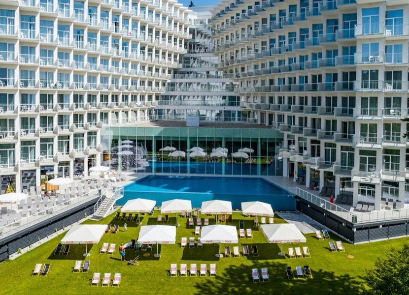 Hotel w kształcie litery U z dużym odkrytym basenem, schodkowymi balkonami, przeszklonym tarasem i trawnikiem z parasolami; goście odpoczywają