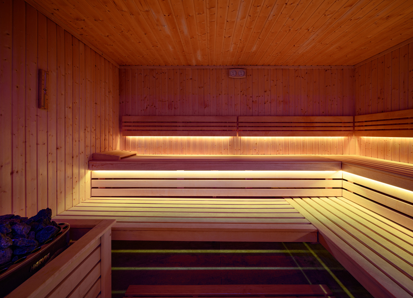 Dřevěná hotelová sauna s vícepodlažními lavicemi, LED podsvícením, kamenným topidlem a nástěnným teploměrem — relax ve spa.