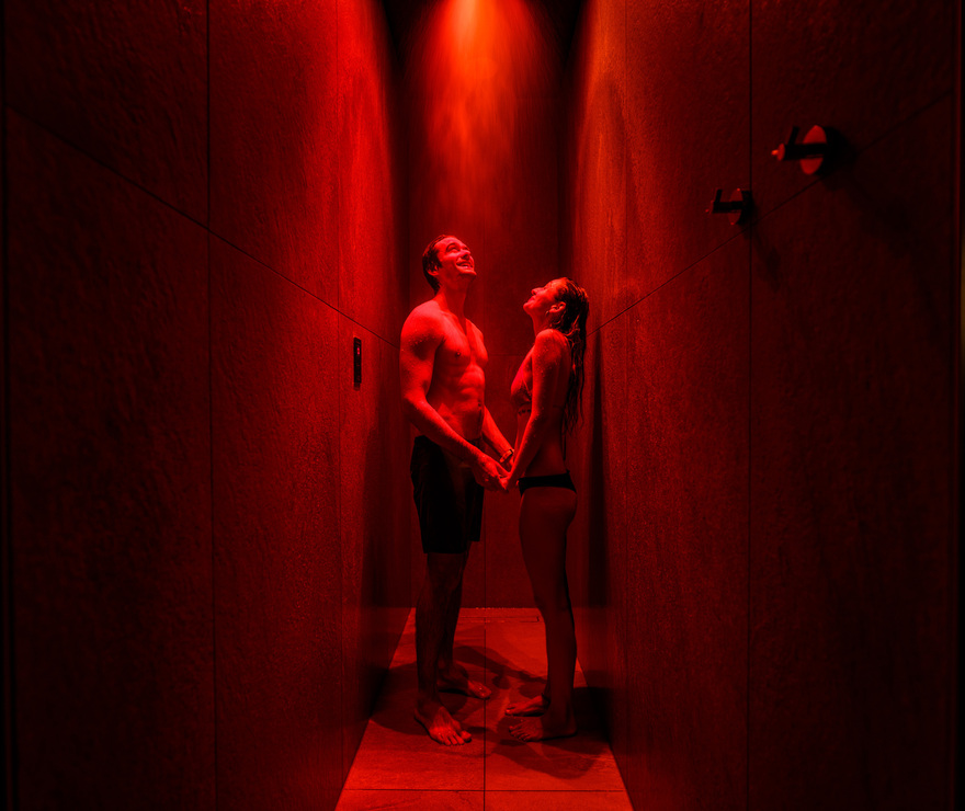Pár drží za ruce pod červeně nasvícenou spa sprchou v úzké obložené místnosti, užívá si chromoterapii.