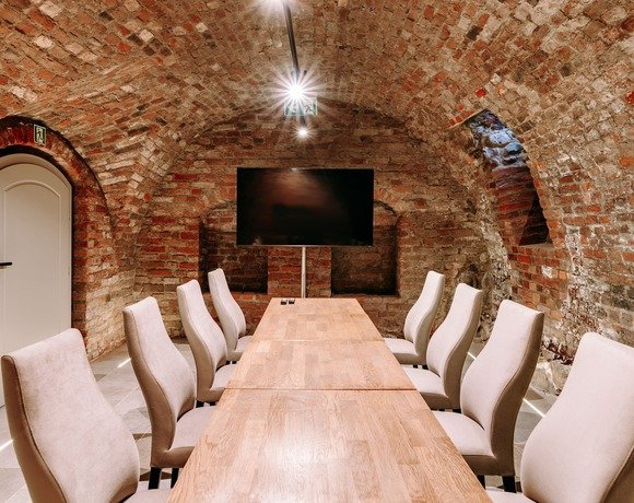 Sala konferencyjna w hotelu z zabytkowymi, ceglanymi sklepieniami i nowoczesnym ekranem TV nad długim stołem i wygodnymi krzesłami.