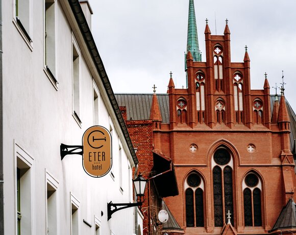 Elegancki hotel Eter z białą fasadą na tle czerwonej gotyckiej katedry z wysokimi oknami i zielonymi wieżami