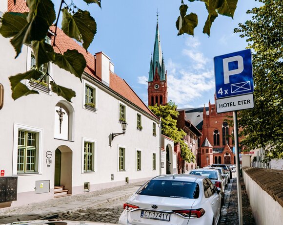 Hotel Eter z biało-czerwoną elewacją i zielonymi oknami przy brukowanej ulicy, z widokiem na neogotycki czerwony kościół.