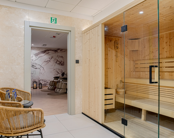 Nowoczesna sauna drewniana z przeszklonymi drzwiami, obok dwie wiklinowe fotele z butelkami wody, widok na pokój relaksu z masażerami.