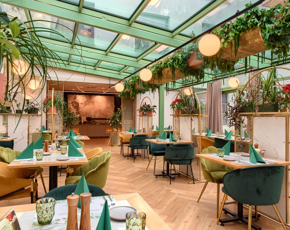 Restauracja z przeszklonym sufitem, zielone i musztardowe wygodne fotele, zielone serwetki, dekoracje roślinne i miękkie oświetlenie.